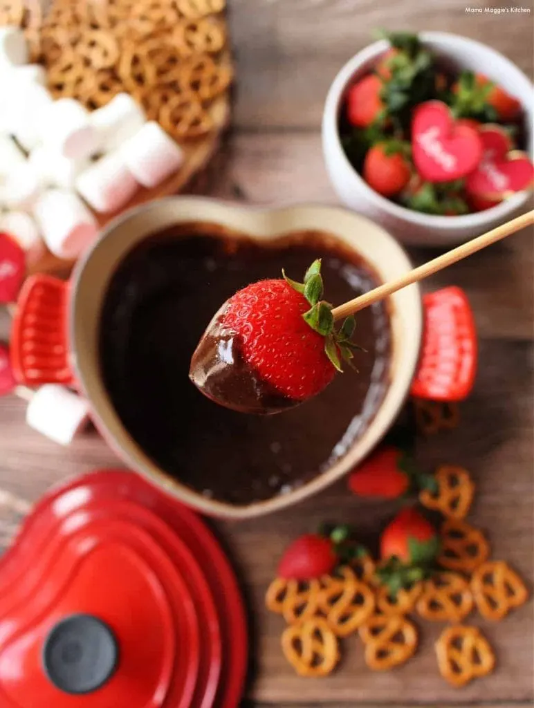 Heart shaped pot of chocolate fondue with strawberry dipped on a stick.