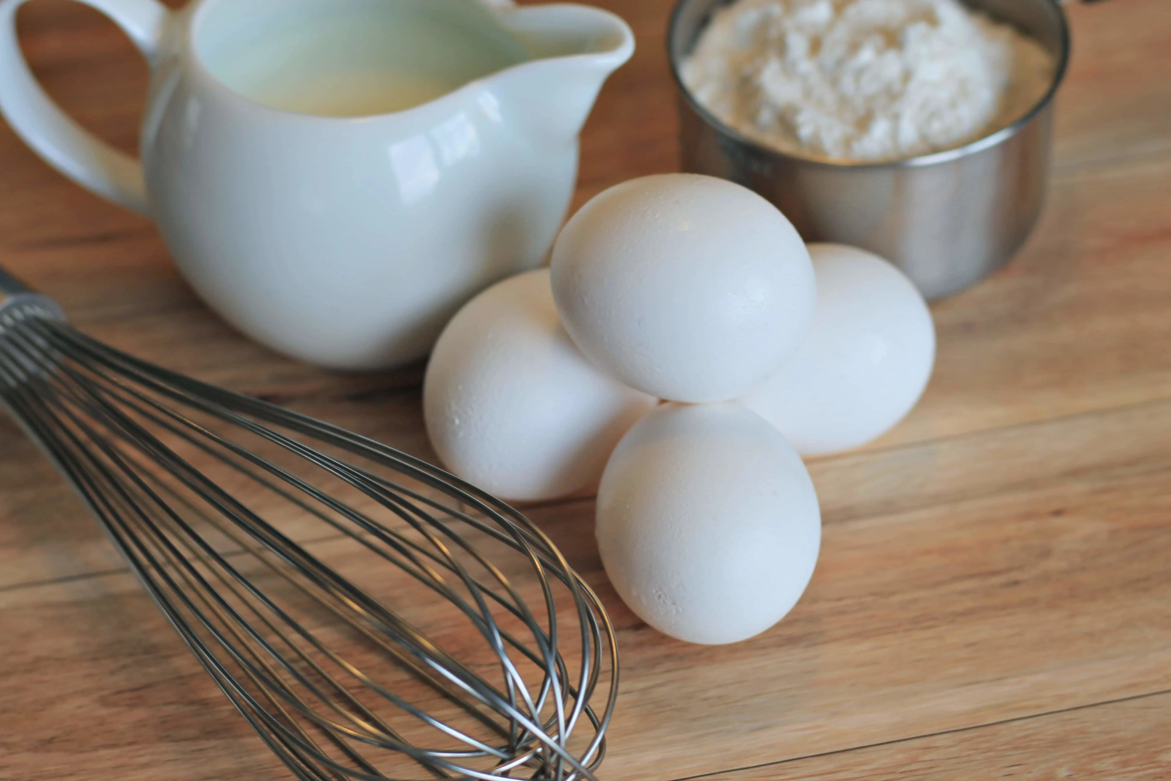 Eggs and flour on wooden cutting board with metal whisk.