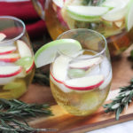 White Wine Sangria in stemless wine glasses with full pitcher behind - fresh fruit and rosemary on the cutting board