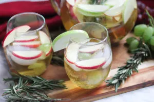 White Wine Sangria in stemless wine glasses with full pitcher behind - fresh fruit and rosemary on the cutting board