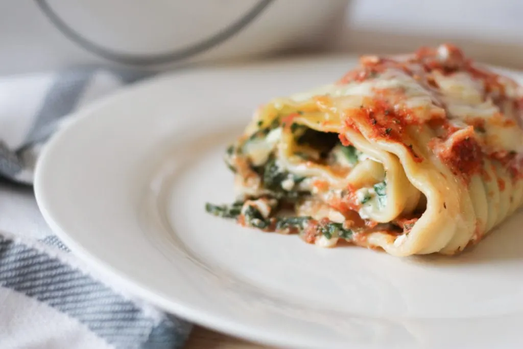 White dinner plate with cooked lasagna roll up set a top a wooden cutting board with tea towel.