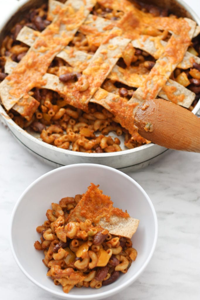 Skillet of Chili Mac with wooden spoon resting on the side and a white bowl with single serving.  