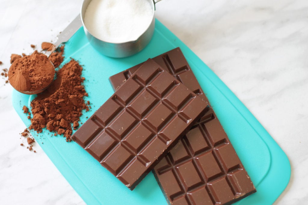 Teal cutting board with scoop of cocoa powder, measuring cup with sugar and two chunks of dark chocolate bar