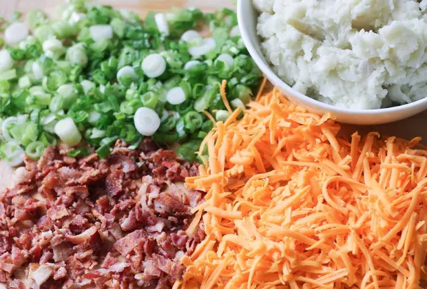 Cutting board with ingredients for casserole - grated cheese, mashed potatoes, crumbled bacon and sliced green onions