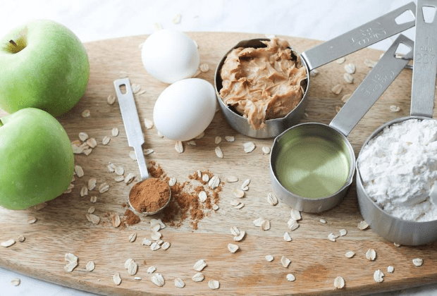 Oval wood cutting board with apples, peanut butter, eggs, flour, oil and spices for making muffins