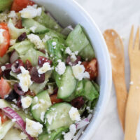 White bowl filled with Greek salad on marble countertop with wooden utensils.