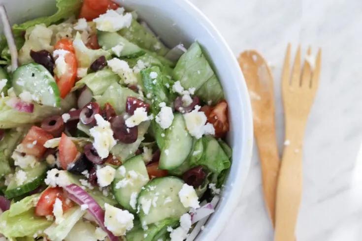 White bowl filled with Greek salad on marble countertop with wooden utensils.