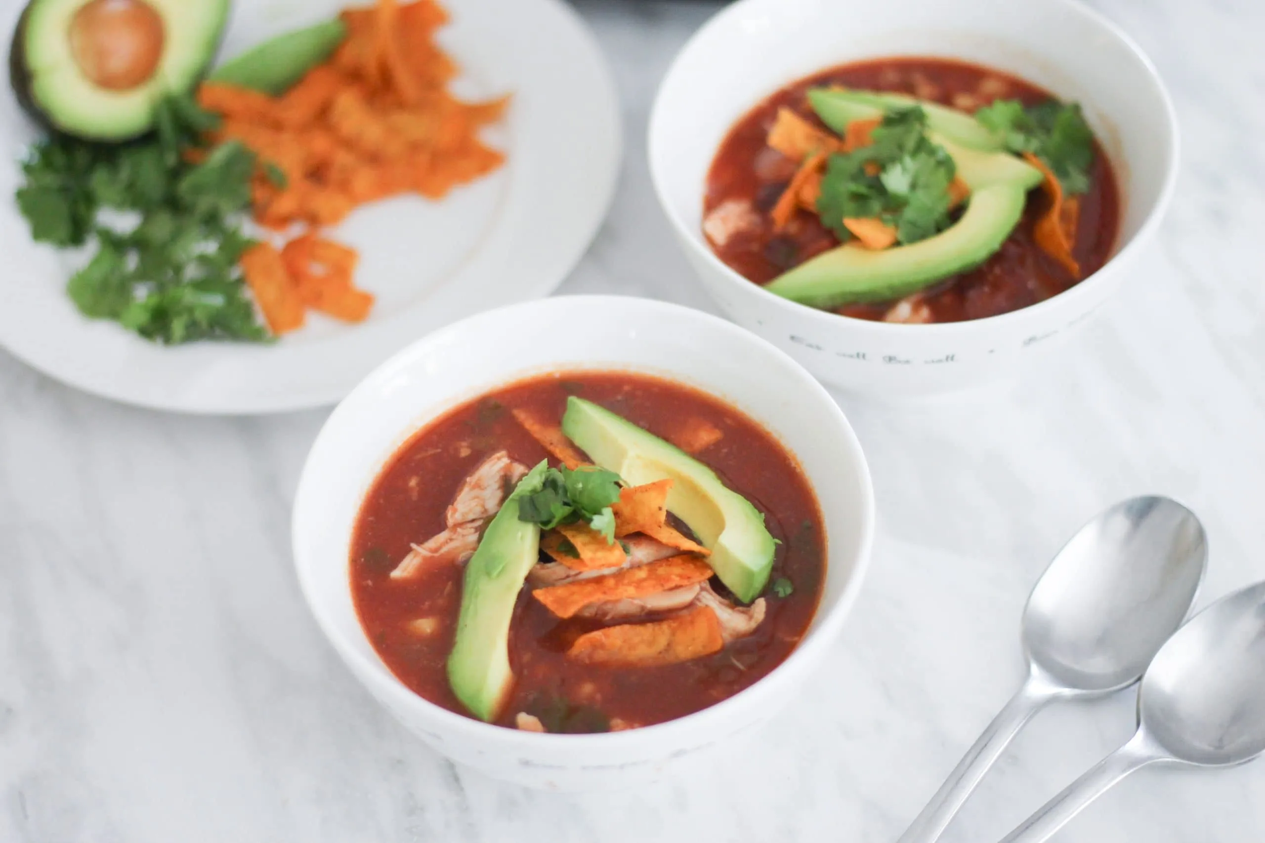 White bowls filled with chicken tortilla soup with toppings on a white plate next to it.